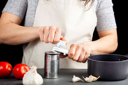 A woman is carefully opening a can of tomato paste on a kitchen counter using a white plastic can opener. She is preparing a meal for which she uses both fresh and pureed preserved tomatoesの写真素材