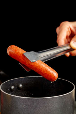 A caucasian woman is lifting up a single hot dog out of boiling water using a metal tong. A nonstick cooking pan and black background  is seen. Homemade cooking, convenient fast food concepts.の写真素材