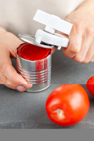 A woman is carefully opening a can of tomato paste on a kitchen counter using a white plastic can...