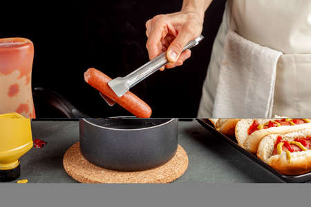 A caucasian woman is taking a fresh boiled hot dog sausage from a pot  using metal tong in order to place it into a hot dog bun. Hot pot is on a cork trivet on stone couter. Ketchup and mustard nearbyの写真素材