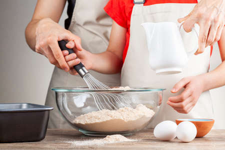 A mother is teaching her little daughter how to cook. They both wear aprons and together they mix a...