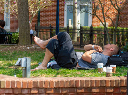 Frederick, Maryland, USA 04-07-2021: A young homeless man wearing hospital bracelet is basking on the grass at a local park in Frederick. He is bare feet, lying crosslegged and shaving his beard.のeditorial素材