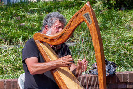 Frederick, MD, USA 04-07-2021:  Closeup image of a street musician performing on the sidewalk by playing his harp. The busker sits on a foldable chair in Carroll Creek Park downtown, Frederickのeditorial素材