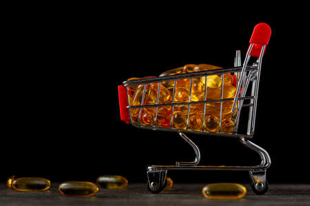 Dark background image with a toy shopping cart filled with omega-3 fish oil pills. A concept image for purchasing over the counter dietary supplements and the market for these items.の写真素材
