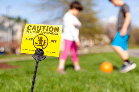 A yellow yard sign warning kids and pets of the recent pesticide spraying and advices them to stay away. Kids are playing soccer in the background regardless. Pesticide use is a big concern.の写真素材