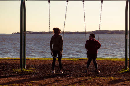 04-16-2021 Oxford, MD, USA: A young couple is sitting on swings at a playground overlooking the waterfront park by Tred Avon River and they are watching the sunset from the prime location as they chatのeditorial素材