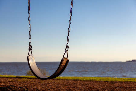Abstract concept showing close up   empty swing seat with the chains overlooking river or sea at sunset. Loneliness, being alone, romance, childhood, moody, pandemic, lockdown and child themes.の写真素材
