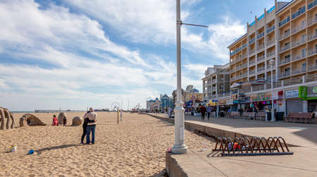 04-18-2021 Ocean City, MD, USA: A young couple is hugging on the sandy beach of Ocean City, Maryland on a lovely afternoon. In the background, there is board walk, pier, fun fair, buildings.のeditorial素材