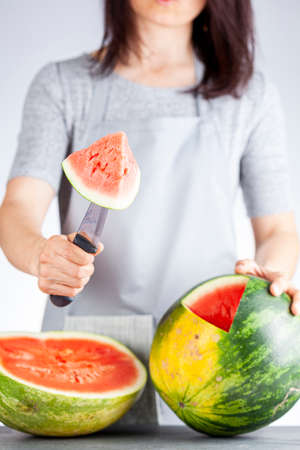A young caucasian woman is cutting a wedge out of a ripe watermelon using a knife. Melon is ripe with field spot. Light and airy summer season fruit concept for refreshing raw healthy fruit dietの写真素材