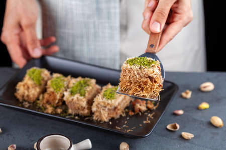 A Turkish woman is serving a slice of delicious traditional kadayif dessert that is  made with shredded phyllo dough mixed with walnuts and ground pistachio topping. A syrup pitcher is for sweetening.の写真素材