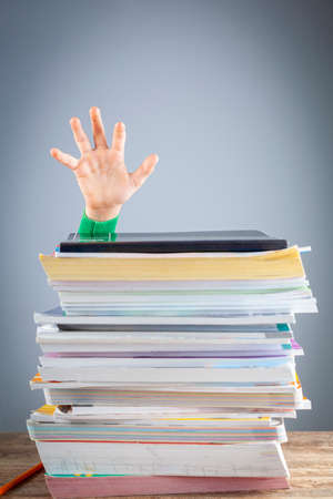 Abstract concept image showing a young student behind a large pile of test prep books on a study desk. An overwhelming load. The kid is trying to raise hand. A demanding curriculumの写真素材