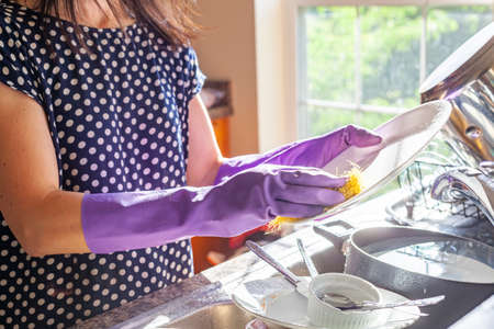 A woman wearing purple work gloves is doing dish washing. She is scrubbing a white porcelain plate using detergent and a sponge. Gloves protect hands from adverse effects of detergents.の写真素材