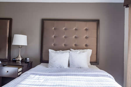 Closeup image of a ready made queen size bed with white bedding and a bunch of pillows. An upholstered head board is on the wall. A generic hotel room interior with night stand, drawers and lamp.のeditorial素材