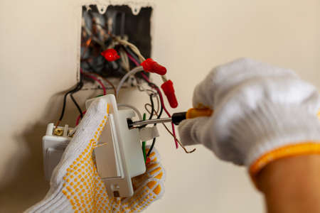 An electrician is replacing a wall switch. A DIY project concept. High voltage danger. installing wire connection using screw driver. The professional wears protective rubber gloves for safetyの写真素材