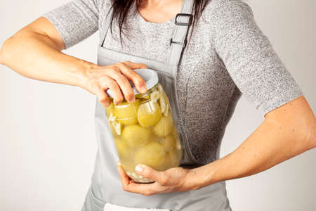 A caucasian woman chef wearing apron is trying to open a stubborn jar lid. She uses force to unscrew the lid in kitchen. Concept image for pickling tomatoes and gherkins and difficulty in opening lidsの写真素材