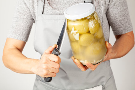 A caucasian woman wearing apron is trying to open a stubborn lid. She is inserting a sharp kitchen knife between the jar and the lid to relieve pressure and unscrew the tight lid.の写真素材
