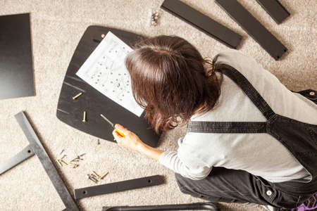 DIY furniture assembly concept including a caucasian person sitting on carpet floor with pieces of furniture, trying to assemble it based on instructions. She holds instruction sheet and a screwdriverの写真素材