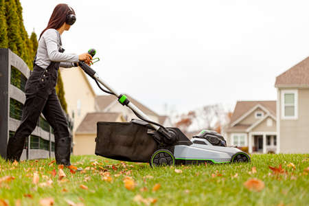 a young caucasian woman wearing overalls and noise blockers is operating a lawnmover on grass in late autumn. There are fallen leaves and a suburban neighborhood setting in background.の写真素材