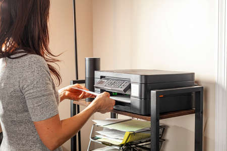 Concept image for office work. A woman is placing or removing paper. There is a wireless photocopier, scanner, printer combo device on top of a shelving unit at a small office setting.の写真素材
