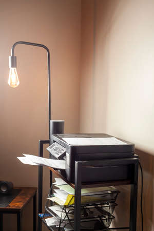 Small home office or cubical with a floor lamp, a bookcase and printer scanner on top of it. There are multiple folder and papers in organisers in the shelves. Working from home concept.の写真素材