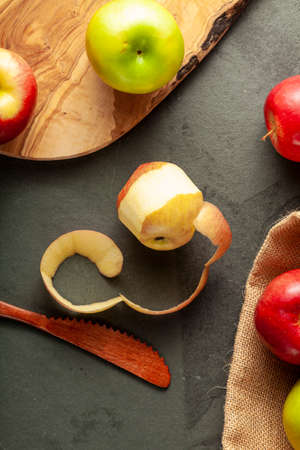 apples of different varieties are seen on dark stone and wooden background. still life image with half peeled apple and wooden knife. Versatile concept imageの写真素材