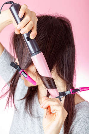 A beautiful young caucasian woman is using a conical electrical curler wand or curling iron to create curles in her red brown hair. DIY cosmetics concept against pink background.の写真素材