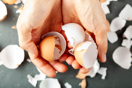 A woman is holding various sizes and shapes of white and brown empty eggshells in her hand. Pieces of broken eggs are seen in background. Flat lay concept image.の写真素材