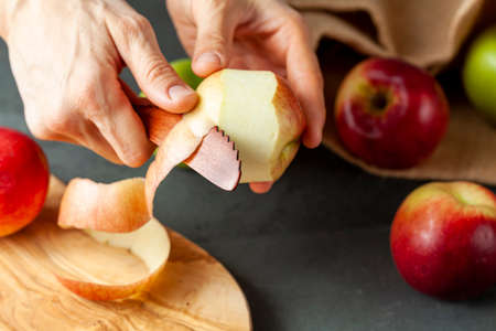 apples of different varieties are seen on dark stone and wooden background.  A woman is peeling the skin of an apple using a fruit knife. Healthy eating and cooking conceptの写真素材