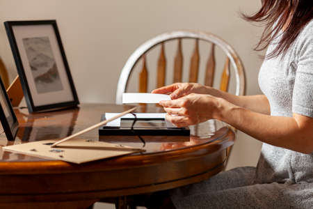 A young caucasian woman is placing a printed photograph into a picture frame with kickstand. There are other frames on the table. Tabletop picture framing concept.の写真素材