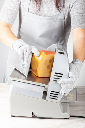 A chef is slicing a large chunk of swiss cheese using an electrical deli slicer. She wears protective cut resistant gloves while operating the machine.の写真素材