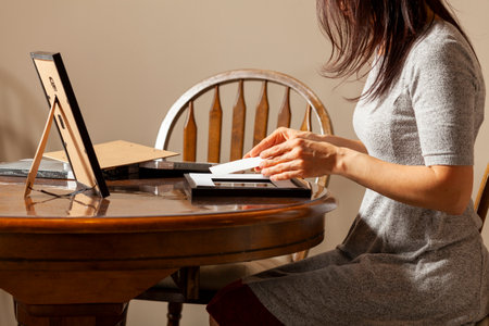 A young caucasian woman is placing a printed photograph into a picture frame with kickstand. There are other frames on the table. Tabletop picture framing concept.の写真素材