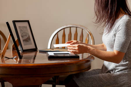 A young caucasian woman is placing a printed photograph into a picture frame with kickstand. There are other frames on the table. Tabletop picture framing concept.の写真素材