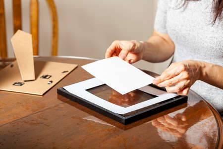 A young caucasian woman is placing a printed photograph into a picture frame with kickstand. There are other frames on the table. Tabletop picture framing concept.の写真素材