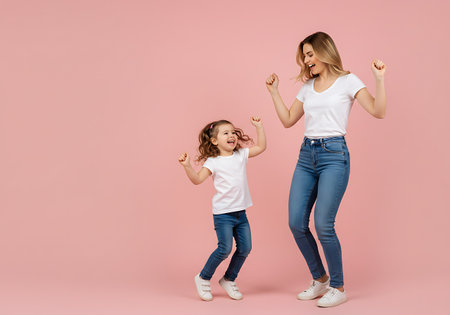 A happy mother and her young daughter in white t-shirts and jeans dance with excitement against a plain pink background.の素材