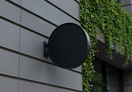 A blank, dark circular sign is attached to a gray paneled wall, with lush green plants cascading nearby.の素材