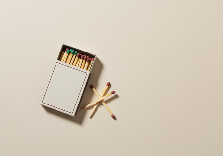A white cardboard box of matches is open, revealing colorful heads. Several wooden matchsticks are scattered nearby on a neutral surface.の素材