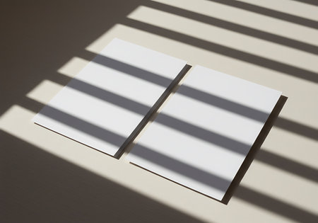 Overhead view of two white notebooks casting shadows, with light filtering through blinds creating a striped pattern.の素材