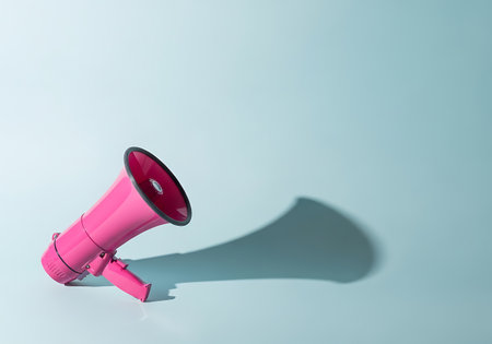 A vibrant pink megaphone lies on a smooth, pastel blue surface, its elongated shadow stretching across the frame.の素材