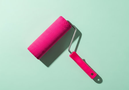 A bright pink paint roller with a metal frame and handle rests on a smooth, light teal surface, casting a subtle shadow.の素材