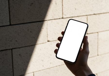 A person's hand grips a sleek mobile device displaying a white, empty screen against a textured wall.の素材
