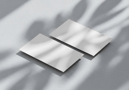 Two blank white business cards are artfully arranged on a textured surface, bathed in dappled sunlight and shadow.の素材