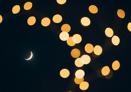 A delicate crescent moon hangs in the dark night sky, surrounded by blurred golden bokeh lights.の素材