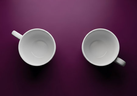 Two empty white ceramic cups are positioned opposite each other on a rich, dark purple surface.の素材