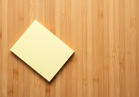 A single, empty yellow note rests diagonally on a warm, grainy wooden background, ready for a message.の素材