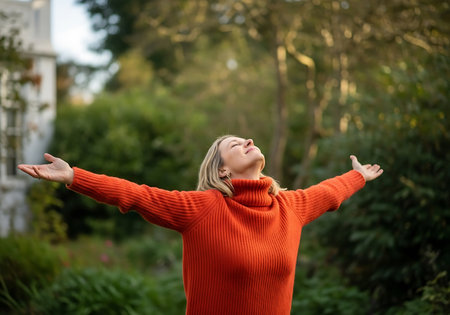 A woman with blonde dreadlocks wears an orange sweater and raises her arms to the sky, enjoying the outdoors amidst lush greenery.の素材