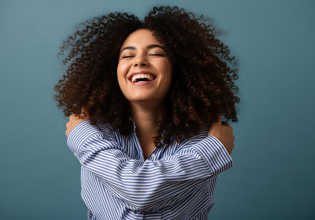A woman with voluminous curly hair laughs heartily, eyes closed, hugging herself tightly against a teal background.の素材