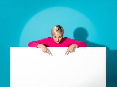 A young woman with blonde hair wears a bright pink top and points down at a large white board. A spotlight effect is visible on the wall.の素材