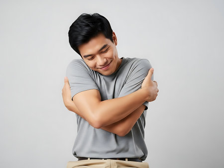 A young man with black hair wearing a gray t-shirt and khaki pants is hugging himself with a happy expression on his face. He is standing against a plain gray background, conveying a sense of contentment and self-love.の素材