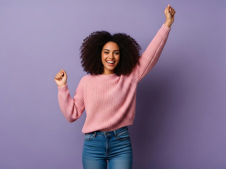 A young woman with curly hair is standing against a purple background, wearing a pink sweater and blue jeans, with her arms raised in celebration.の素材