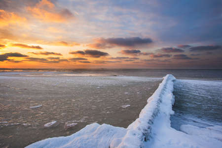 snow-covered breakwaters on a winter beach near a cold, ice-covered sea during sunsetの写真素材